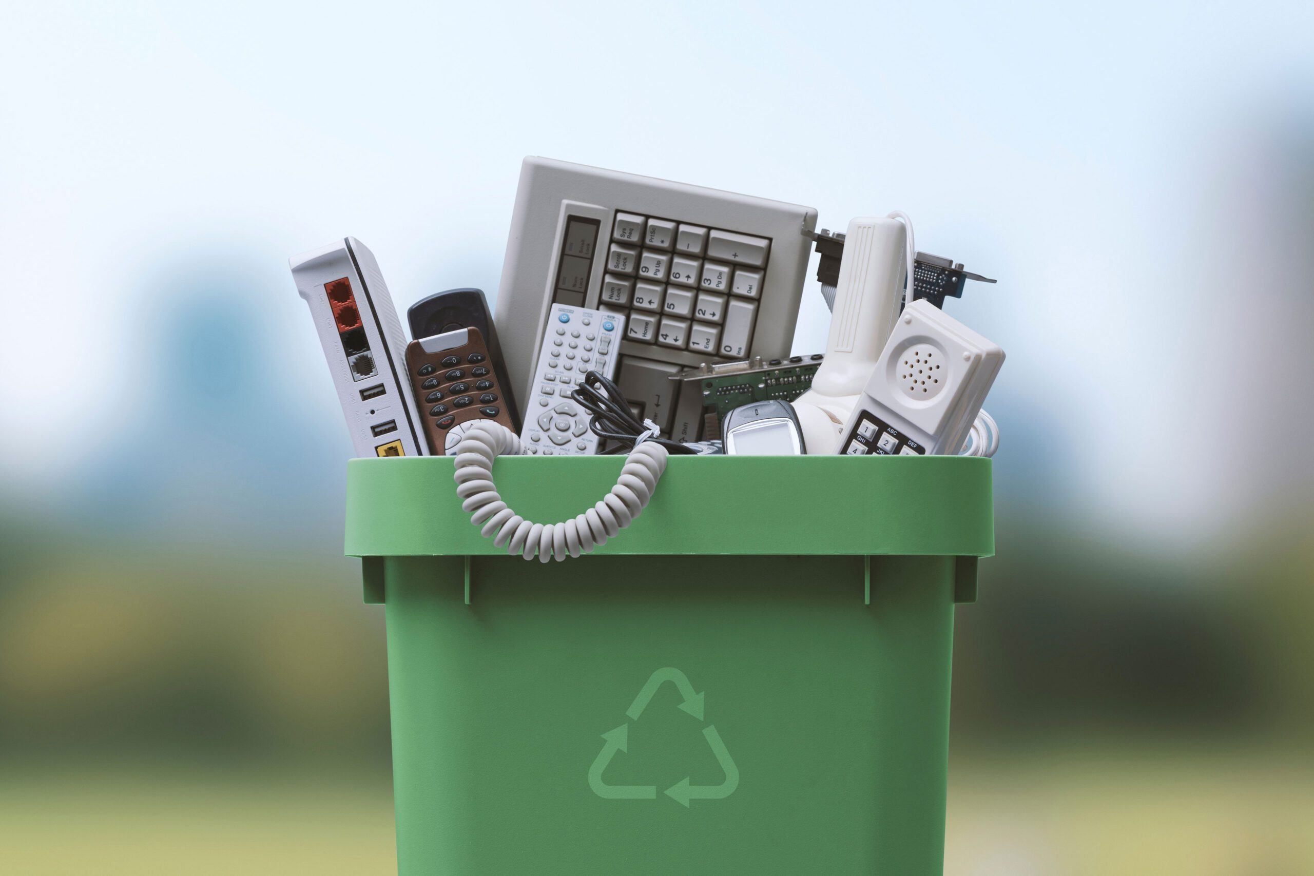 A green recycling bin filled with discarded electronic devices, such as phones, keyboards, remotes, and cables, representing eco-friendly electronics disposal services in Riga, Latvia. Services include secure data wiping, recycling, and fair compensation for salvageable components.